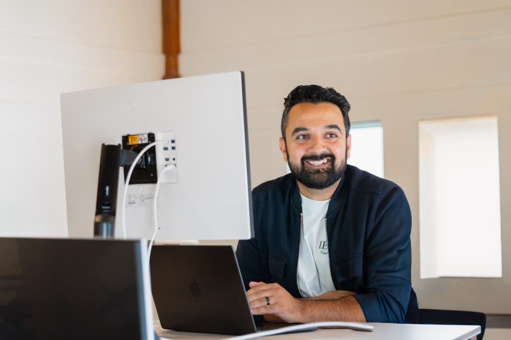 A Gumlooper smiling at a desk beside a monitor and laptop